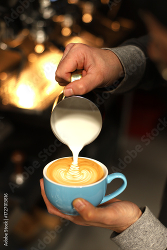 A barista makes a cappuccino with beautiful latte art against the backdrop of a coffee machine with bright background lighting.