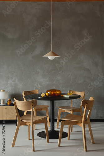 Modern dining area with wooden chairs, round black table, pendant lamp and minimalist decor against textured concrete wall.