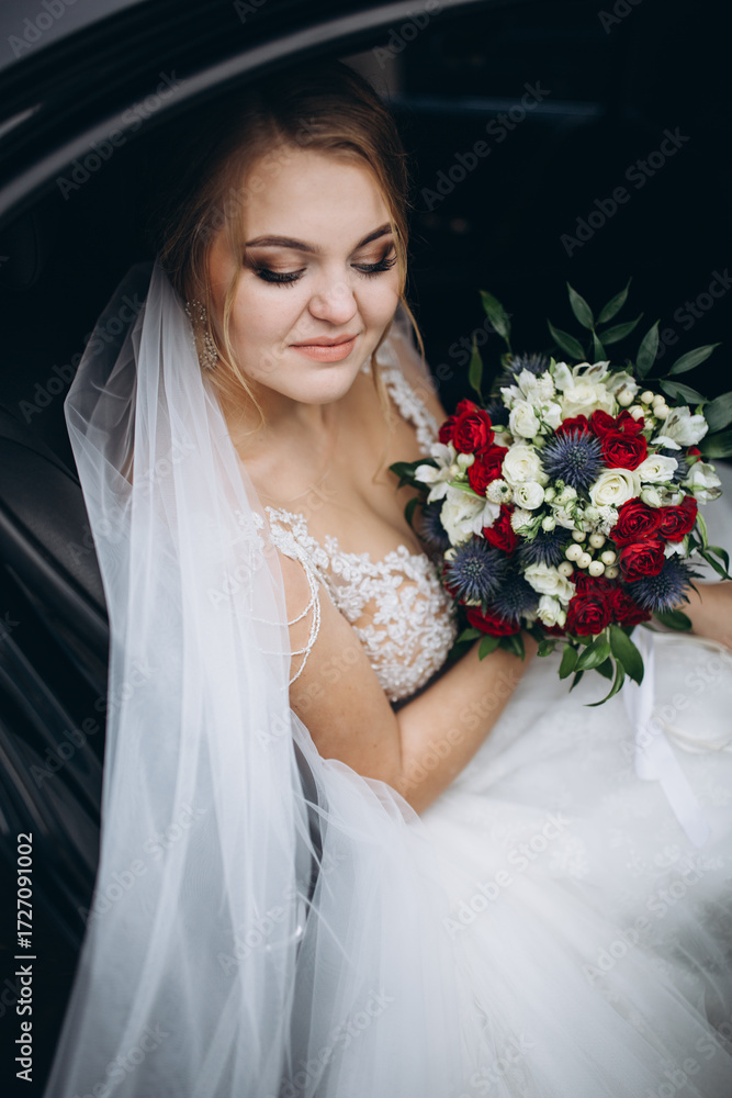 Naklejka premium Beautiful bride in lace wedding dress sitting in a car and holding bouquet of red, white and blue flowers. Smiling woman with veil looking out from vehicle on her wedding day. Elegant bridal portrait.