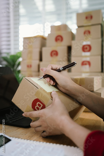 A person checking inventory with a tablet and notebook in a warehouse, surrounded by stacked cardboard boxes for shipping and logistics.