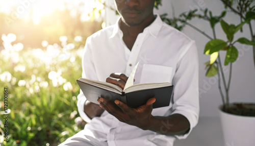 A person in a clean white shirt sits calmly reading a dark-covered book in a sunny garden.