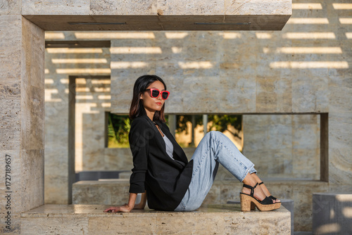 A woman in red sunglasses sits on a stone bench in a modern architectural setting. Spain