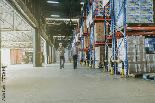 Wide angle view two male warehouse workers moving in a moving aisle area and inspecting the orderliness parcels placed on pallets and shelves in a storage area in an industrial and logistic warehouse.