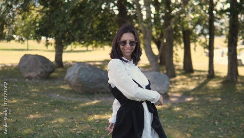 A business woman stands in a park, smiling and wearing sunglasses. She is dressed in a stylish outfit, enjoying the fresh air and greenery around her during a sunny day