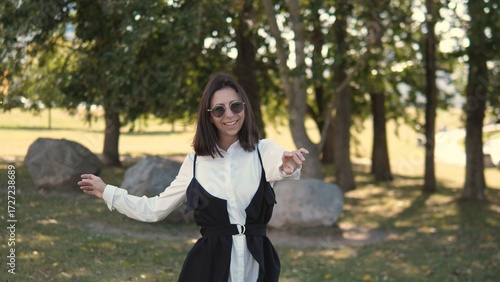 A confident woman dressed in a stylish outfit stands in a park, smiling and enjoying the sunny weather. She is surrounded by greenery and large stones while showcasing her joyful personality