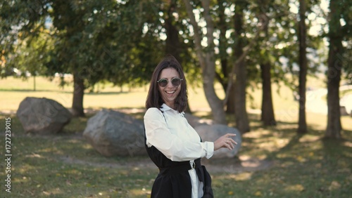 A cheerful businesswoman stands in a park surrounded by trees and rocks. She smiles confidently while wearing stylish sunglasses and a fashionable outfit on a bright day