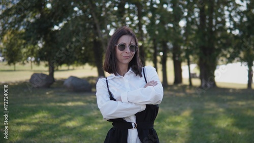 A woman dressed in a black and white outfit stands with her arms crossed in a green park. She wears sunglasses and smiles slightly, surrounded by trees and sunlight