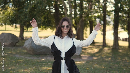 A business woman smiles happily while standing in a park on a sunny day. She wears a chic outfit with stylish sunglasses and is surrounded by greenery and rocks, creating a lively atmosphere