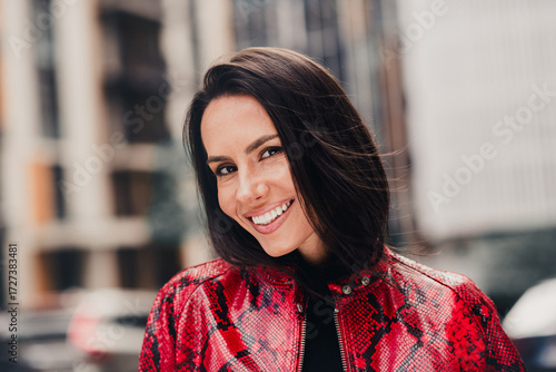 Fashionable brunette woman wearing a stylish red jacket outdoors, smiling brightly in an urban city background on a sunny day