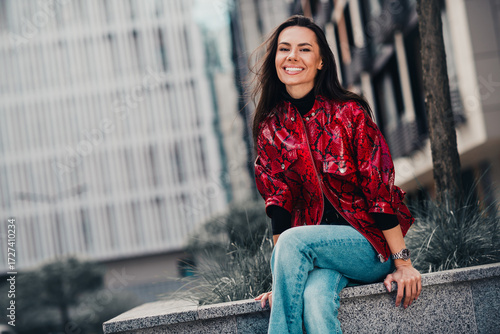 Stylish young woman in red patterned jacket enjoying an urban outdoor setting while smiling confidently and beautifully