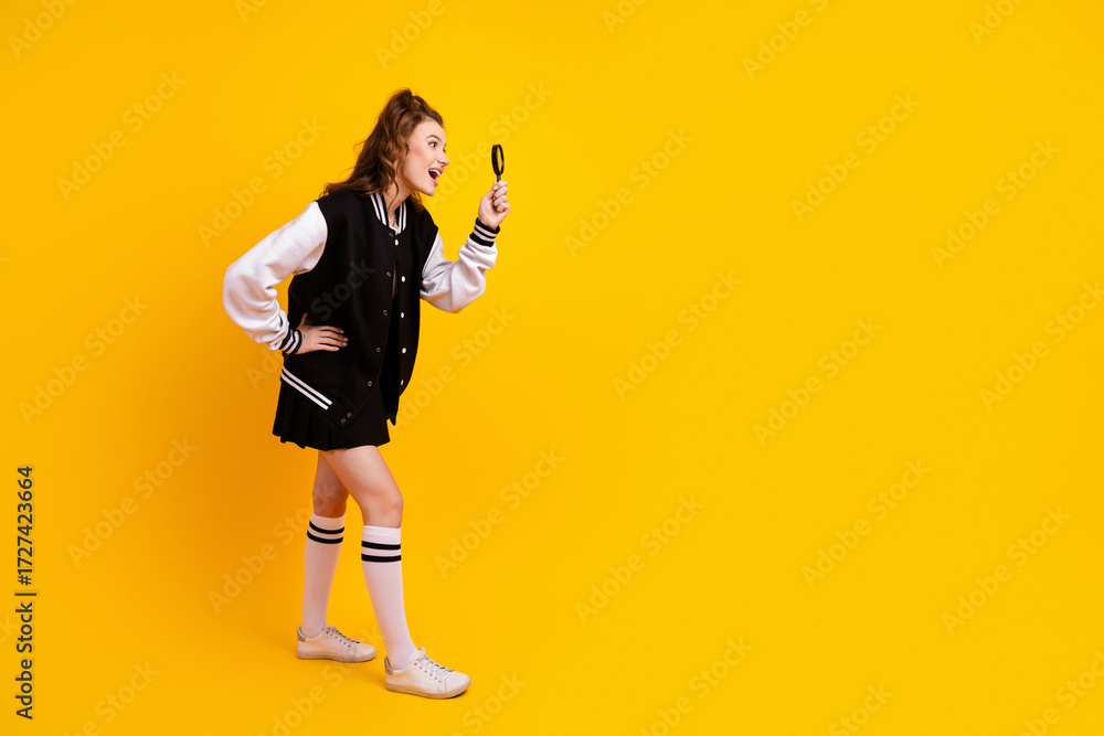 Fototapeta premium Cheerful young student with magnifying glass in trendy jacket posing on bright yellow background, exploring and smiling