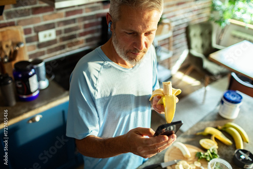 Man eating banana and using smartphone in kitchen