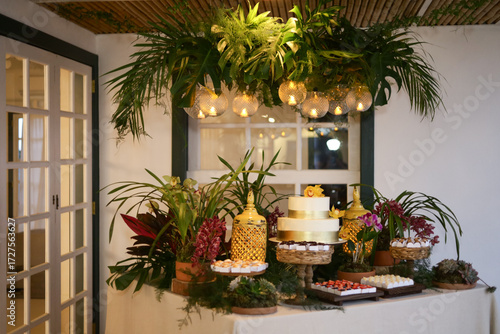 Mesa decorada com doces e flores em ambiente de evento elegante, com iluminação aconchegante e decoração tropical.