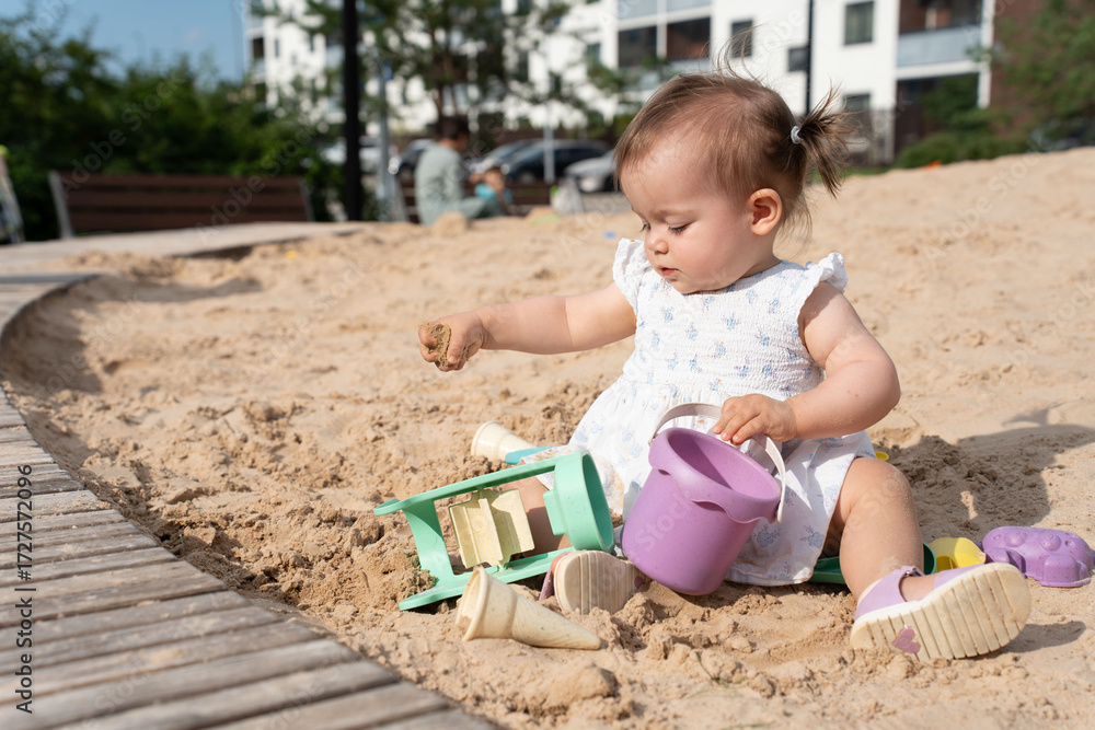 Naklejka premium adorable toddler playing with sand toys on a sunny day in a urban playground surrounded by lush greenery and modern apartment buildings, bliss, interaction, development, relax, peaceful, beige