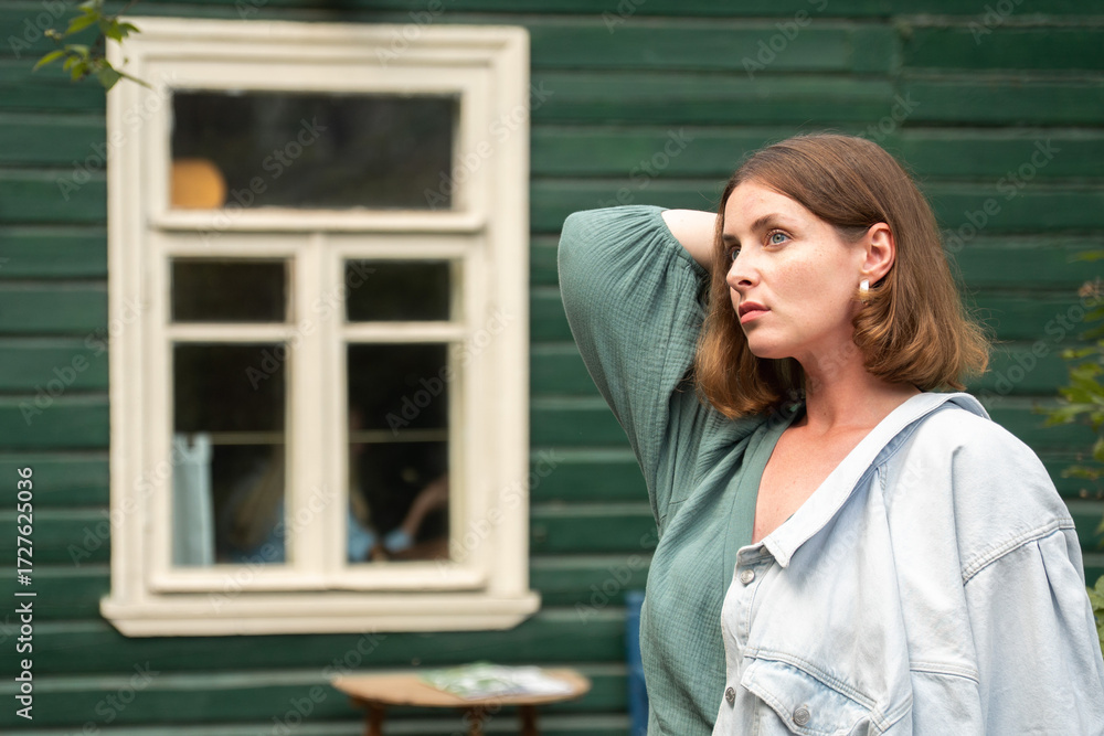 Fototapeta premium A woman in a green dress with a light denim jacket poses near a rustic green wooden house in Peredelkino