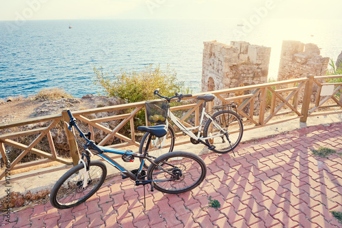 Tablou pe pânză Two Bicycles Parked by Scenic Coastal View With Ruins and Wooden Fencing