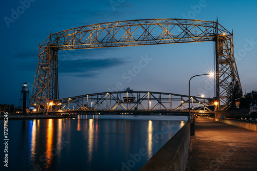 Aerial Lift Bridge illuminated at blue hour over the Duluth Ship Canal in Duluth, Minnesota, with reflections on calm water and the harbor lighthouse in the background.