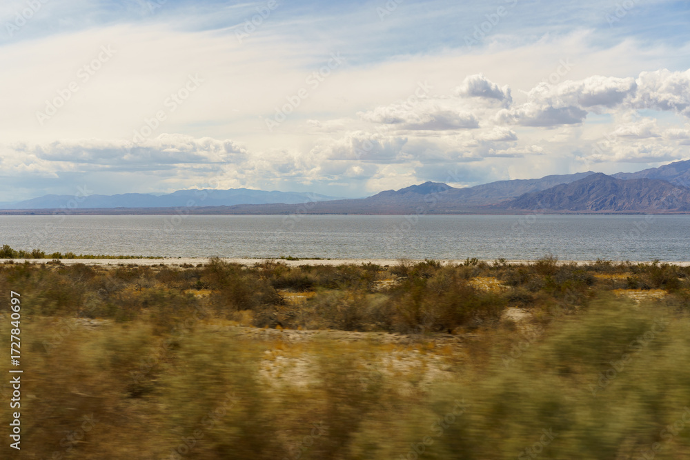 Obraz premium A motion-blurred desert landscape featuring a vast lake and distant mountains under a partly cloudy sky.
