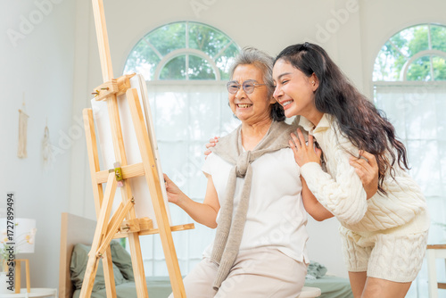 Happy Asian family senior woman and adult daughter drawing and color painting on canvas together in living room. Elderly woman relax and enjoy indoor lifestyle leisure activity and hobbies at home.