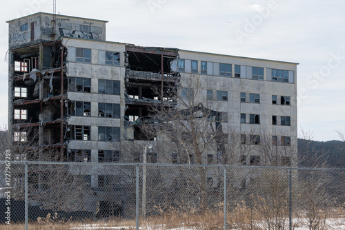 The exterior wall of a desolated large concrete building with broken windows, rusty metal beams, crumbling rubble and debris. The industrial construction site is ruined by an earthquake or bombing. 