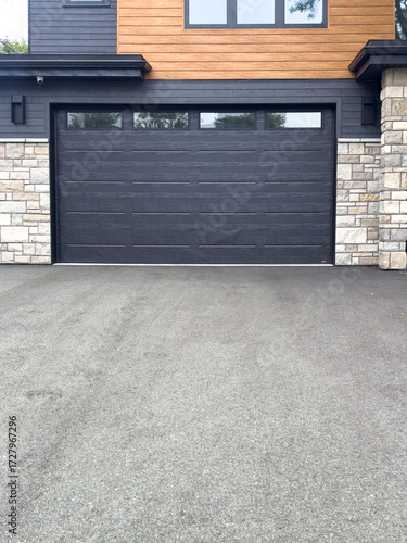 A large black garage door with small glass windows along the top of the wide door. The exterior of the building with beige brick, modern wooden boards, black trim windows, and a modern style entrance.