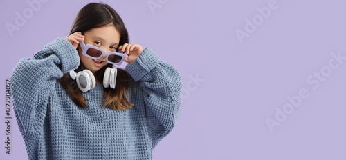 Cute little Asian girl in oversized sweater and with headphones on lilac back...