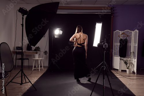 Beautiful young woman with glowing lighting equipment in photo studio, back view