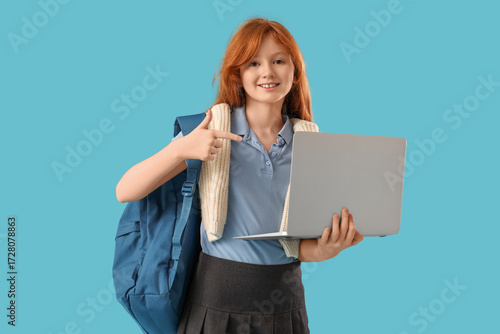Cute schoolgirl with backpack pointing at modern laptop on blue background
