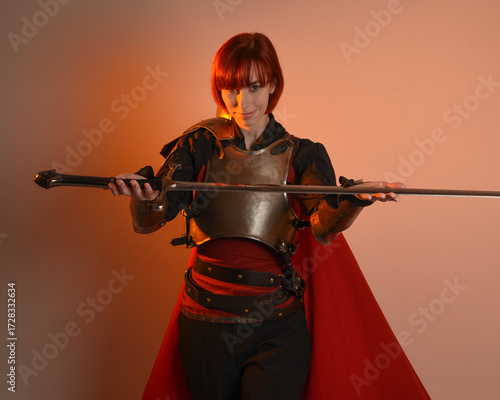 portrait of Fantasy warrior woman model, holding sword weapon, historical medieval armour costume with red fairytale cloak, strong fantasy knight. isolated studio background golden cinematic lighting.