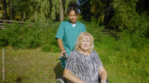 Happy charming senior woman in wheelchair and positive black female carer in uniform looking at scenic summer nature and friendly communicating , while enjoying leisure during stroll outdoors.