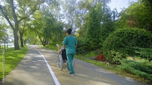 Rear view of African American female caregiver in uniform rolling happy elderly woman in wheelchair on park alley , friendly talking and sharing while enjoying leisure outdoors.