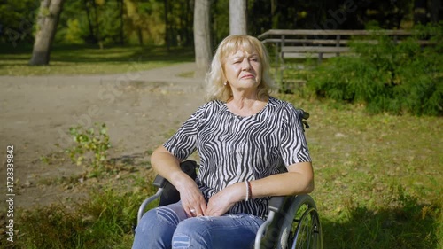Pensive beautiful retired elderly woman with age-related health conditions sitting in wheelchair , looking at scenic nature and thinking about days go by while relaxing in summer nature.