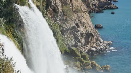 A powerful waterfall cascades into the calm sea. The video captures a stunning natural scene where a large waterfall plunges from a high cliff directly into the blue ocean. 