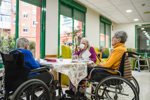 Elderly women in wheelchairs enjoying an activity and drinking coffee in a bright nursing home common area, promoting social engagement and well being