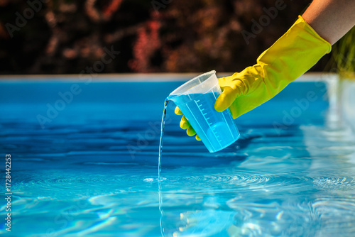 Hand with yellow protective glove pouring blue chemical liquid from measuring cup into swimming pool water for cleaning and disinfection