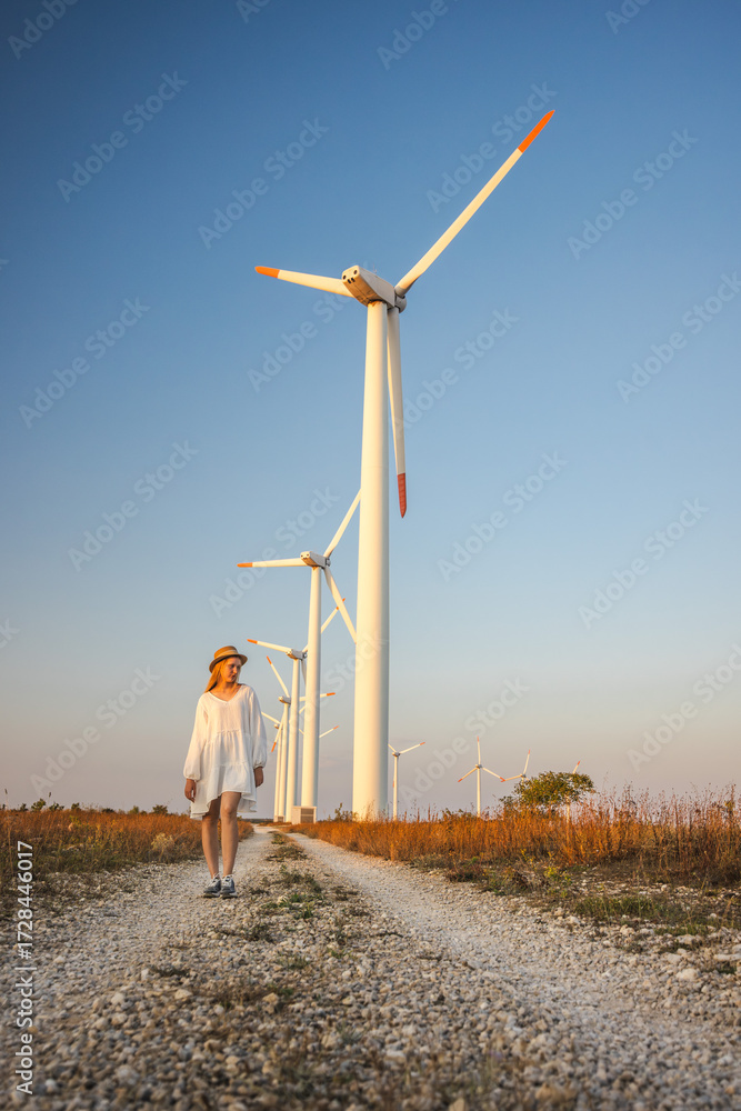 Obraz premium Young woman in white dress and hat walking along a gravel path near wind turbines at sunset.