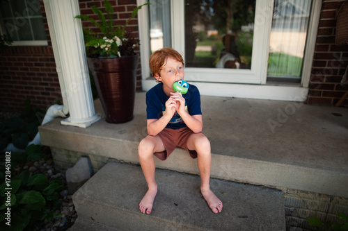 Young boy enjoys cartoon popsicle while sitting barefoot on porch