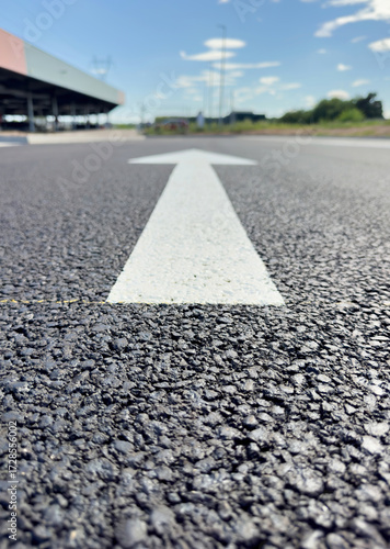 Closeup low angle view of white directional arrow painted on dark asphalt road surface pointing forward with blurred outdoor background sky building