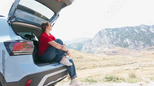 An adult woman enjoys a stunning mountain view while sitting in the trunk of her car