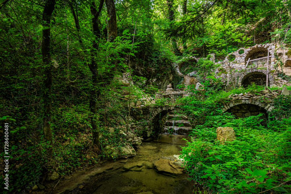 Fototapeta premium A small arched waterfall with a pool, by the old abandoned mill ruins in the forest near the Stomaž village near Ajdovščina, Slovenia