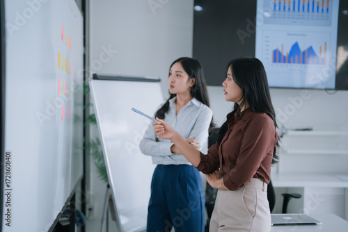 Asian businesswomen collaborating, discussing points, and planning strategies using sticky notes on a whiteboard during a professional office meeting with data displayed on a screen