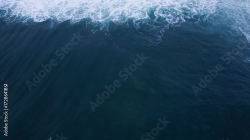 Aerial flying drone top view of huge and slow Atlantic ocean waves crashing over rocky coast at Farol da Nazaré in Nazare, 4K aerial video. Moody February day in Portugal.