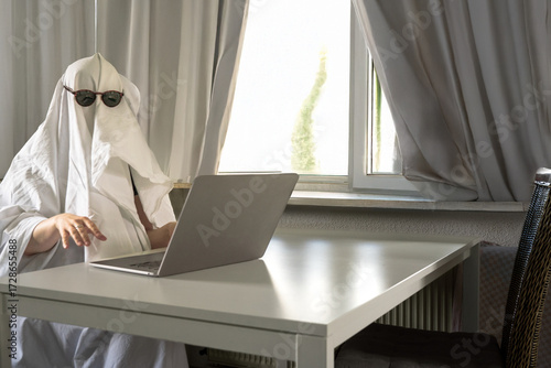 White bed sheet host looking at you while working on a laptop n a spooky grey room