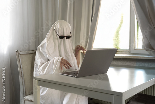 Person in white bedsheet ghost costume working remote on Halloween from home office