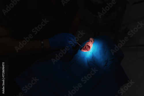 A close-up of a patient during a dental procedure. The mouth is illuminated by a bright blue lamp, while the doctor's blue-gloved hands work with a dental instrument. This emphasizes precision and hyg