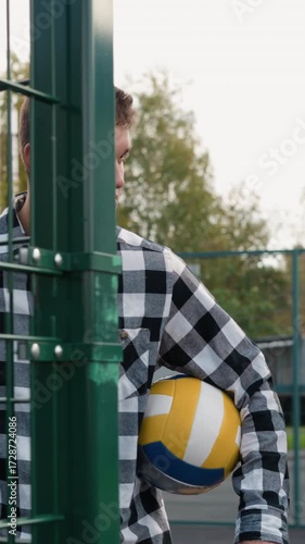 Coach in plaid shirt with a volleyball under his arm opens a green gate, followed by students with a basketball court and trees visible in the background