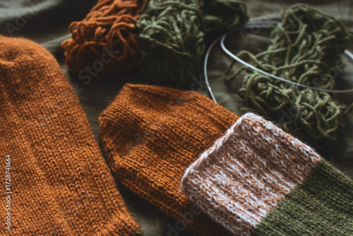 Cozy knitted socks with yarn ball and knitting needles. Striped wool socks on dark green sheet. Cozy autumn colours. Woven crochet close up. Knitting hobby. Knitwear concept. Needlework background. 