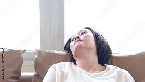 An Asian woman sits on a sofa with a blank expression on her face. She is feeling stressed, sad, and unhappy. She may be experiencing depression.