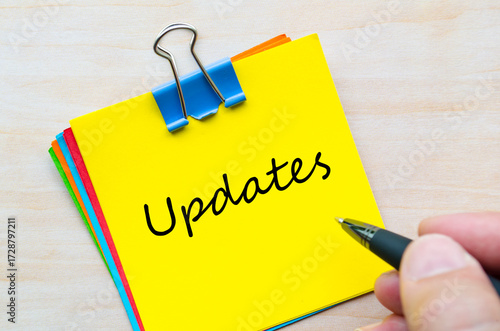 Updates handwritten text on yellow notepad clipped with silver pin on light wooden desk.