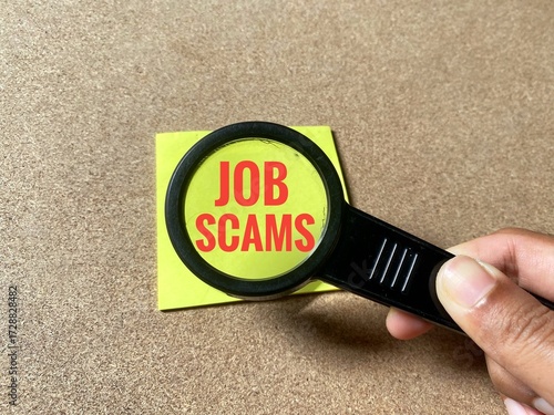 Hand holds a magnifying glass over a yellow sticky note, magnifying the words Job Scams in red text.